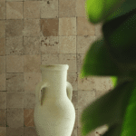 Ściana wyłożona płytkami trawertynowymi Ivory w formacie 10x10 cm, z dekoracyjnym dzbanem na postumencie z kamienia i liśćmi rośliny na pierwszym planie.