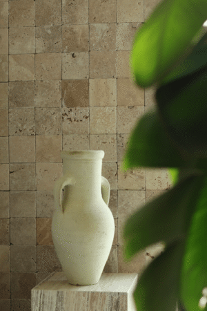 Ściana wyłożona płytkami trawertynowymi Ivory w formacie 10x10 cm, z dekoracyjnym dzbanem na postumencie z kamienia i liśćmi rośliny na pierwszym planie.
